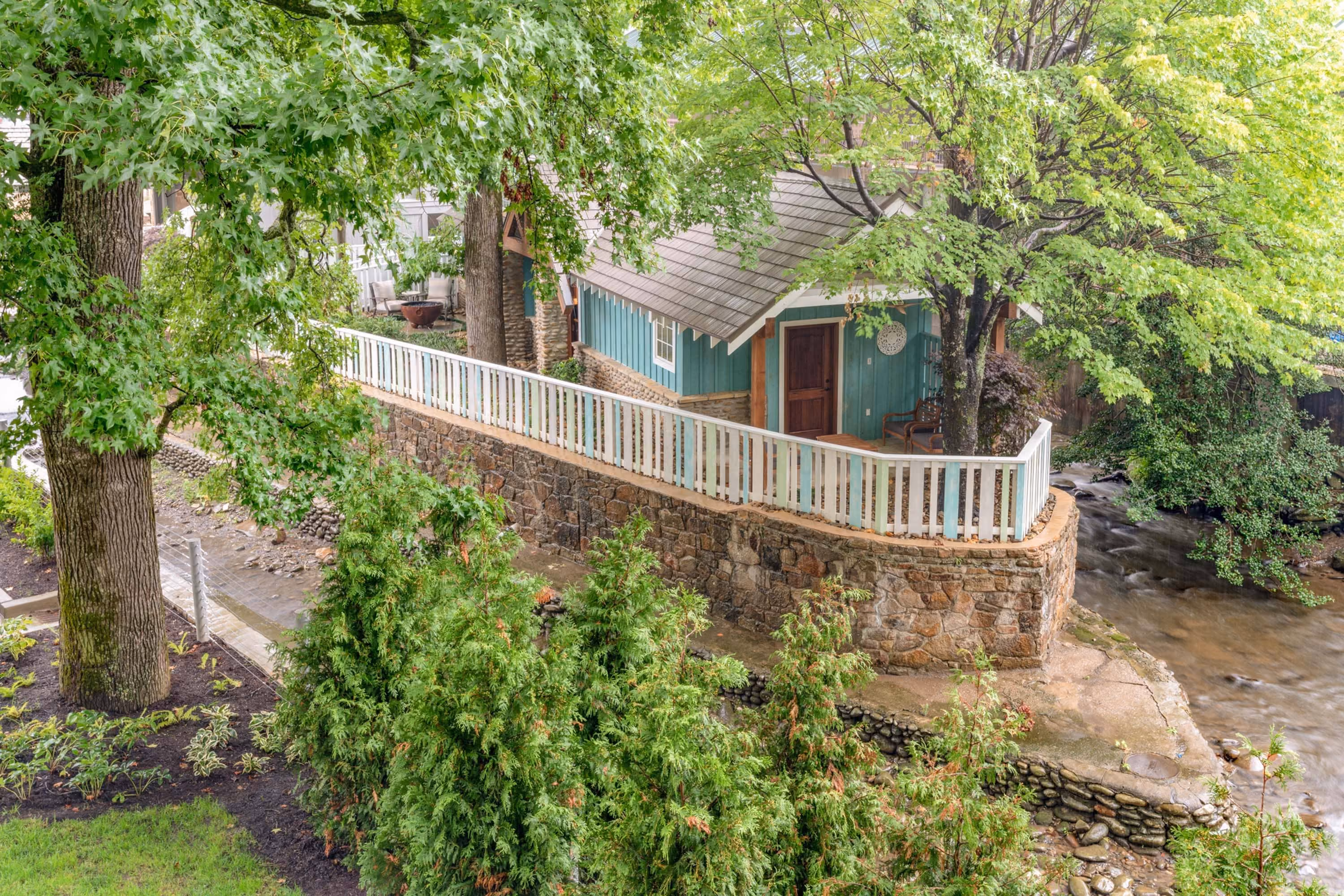 Small teal cabin beside a creek with stone retaining wall and white railing, surrounded by trees
