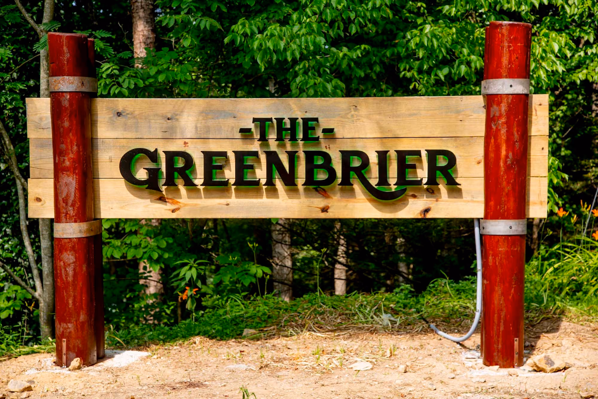 Wooden “The Greenbrier” sign mounted between red posts in a wooded area