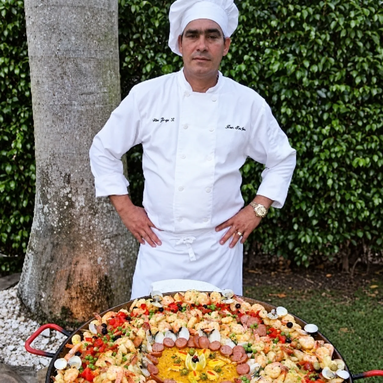 Chef Jorge in a white chef uniform beside a large seafood paella and a palm tree.