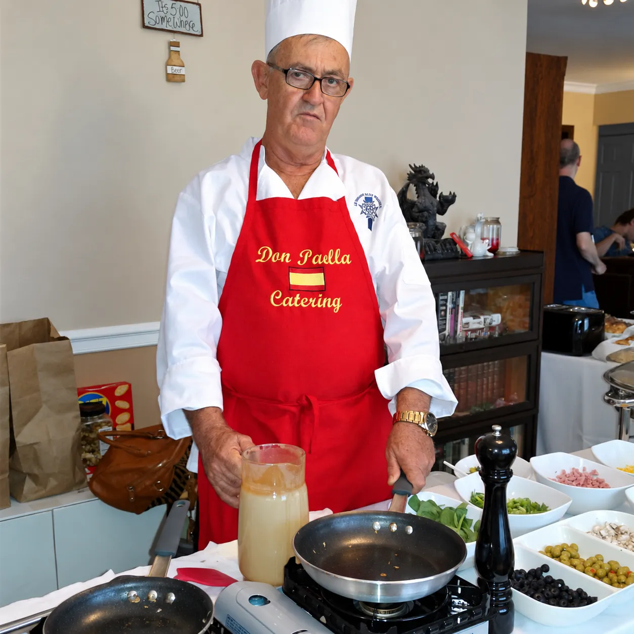Chef Agustin wearing a white toque and red Don Paella apron behind a cooking station.