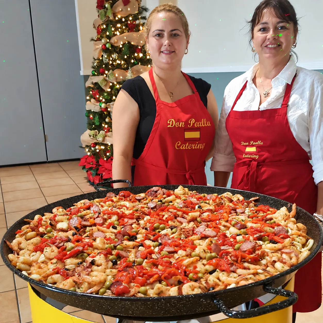 Chefs Islaydis and Geysa in red Don Paella aprons behind a seafood paella beside a Christmas tree.