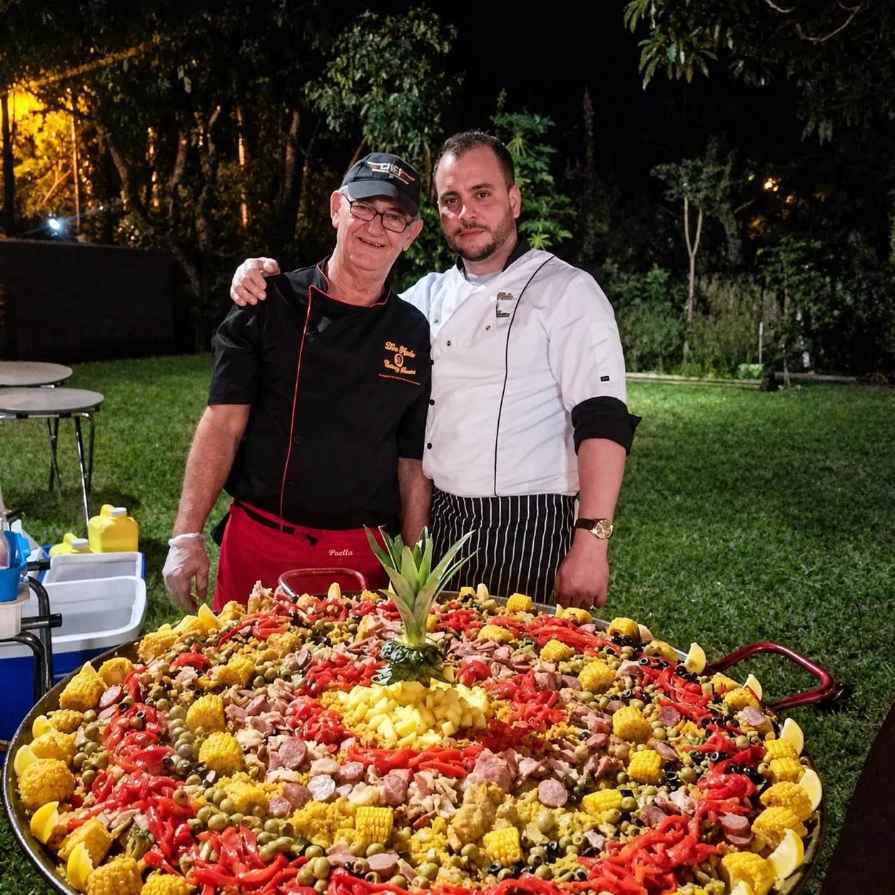Chefs Agustin and Manuel beside a large paella with a pineapple centerpiece at a nighttime garden event.