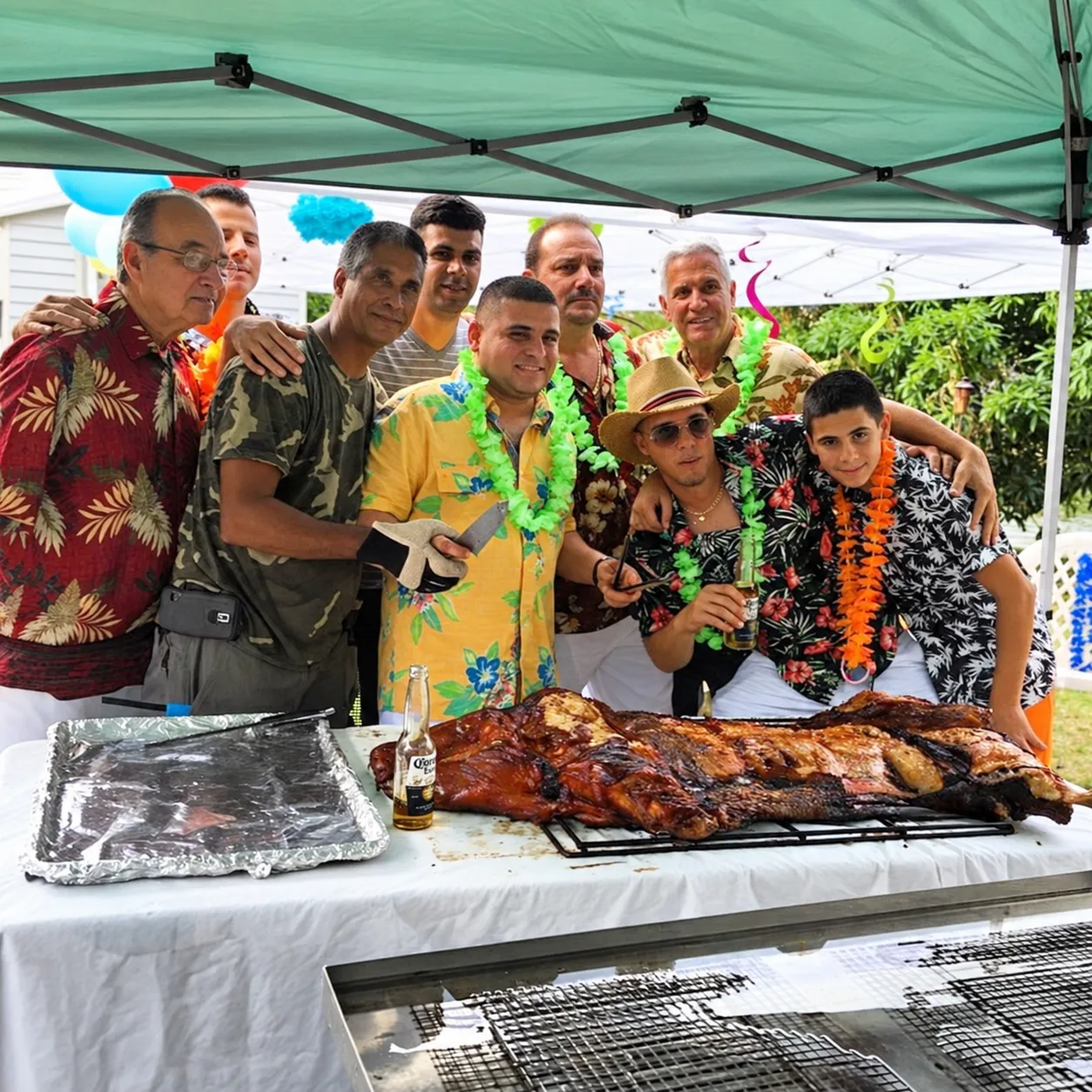 Guests in tropical party attire gathered behind a whole lechón asado under a green canopy