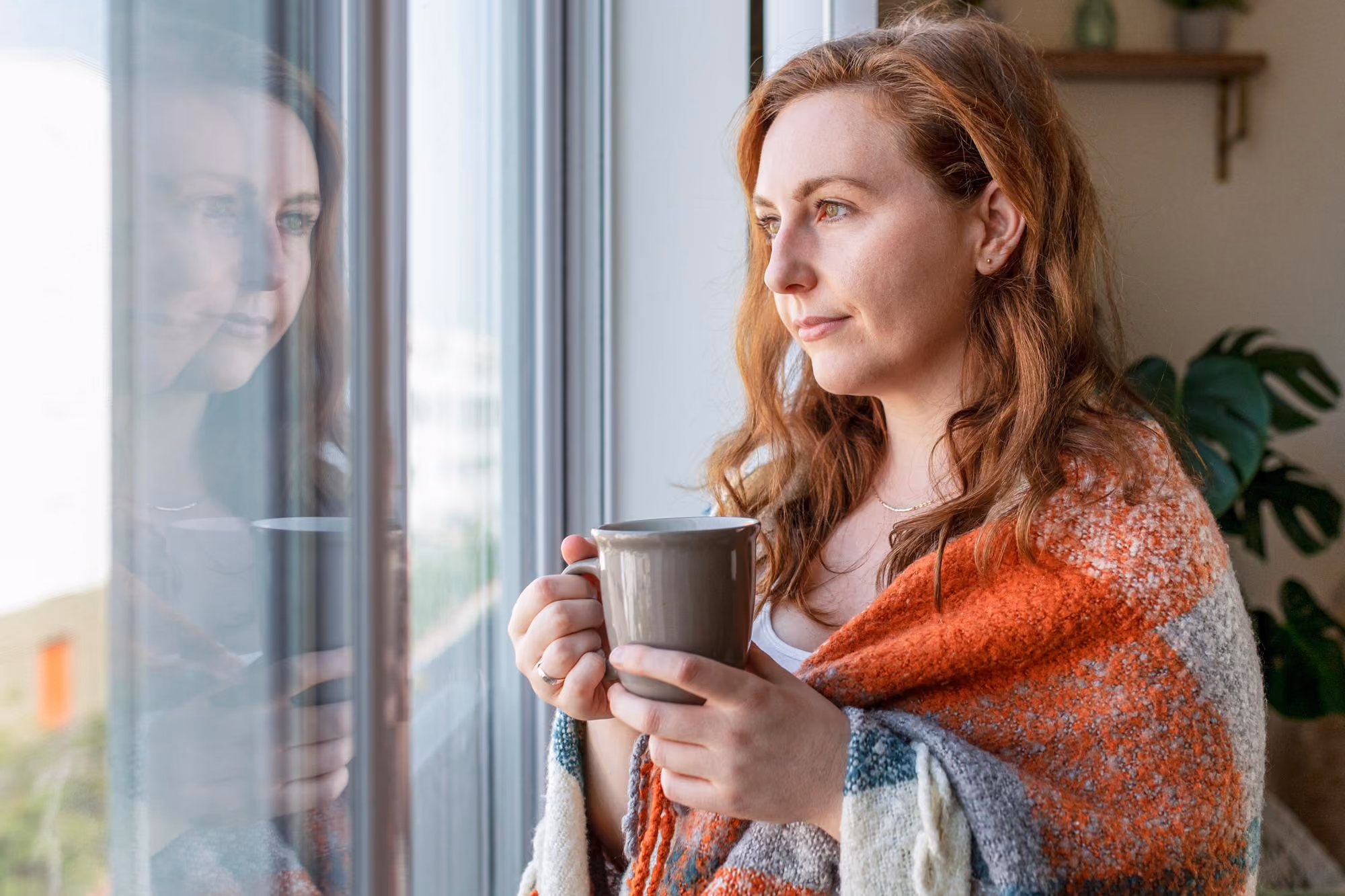 A woman with light brown hair wrapped in a multicolored blanket holds a mug and looks out a window, with her reflection visible in the glass.