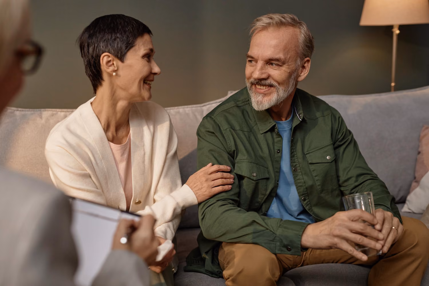 A middle-aged couple sitting on a couch during a conversation with a therapist, the woman with short dark hair and white cardigan smiles while holding the arm of the bearded man in a green jacket who is holding a glass of water and smiling back.