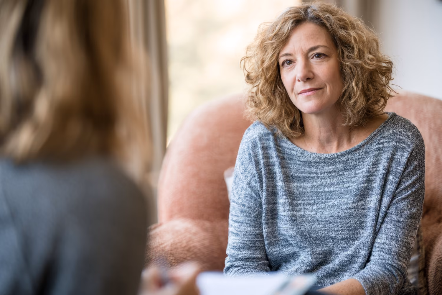 Middle-aged woman with curly hair sitting in a chair, attentively listening to another person whose back is to the camera.