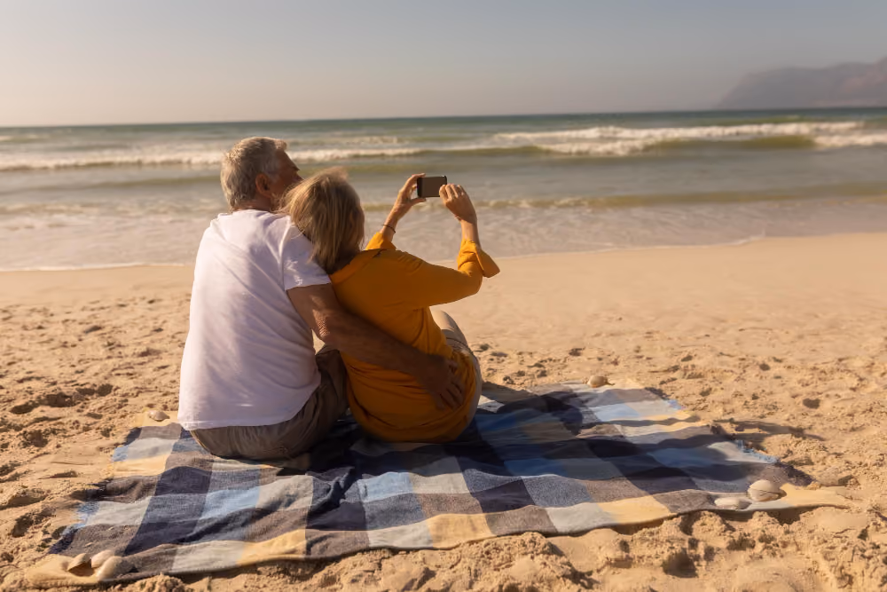 An elderly couple sitting on a blue and beige checkered blanket on the sandy beach facing the ocean, with the woman in a mustard yellow top taking a selfie with her smartphone while the man in a white shirt hugs her from behind.