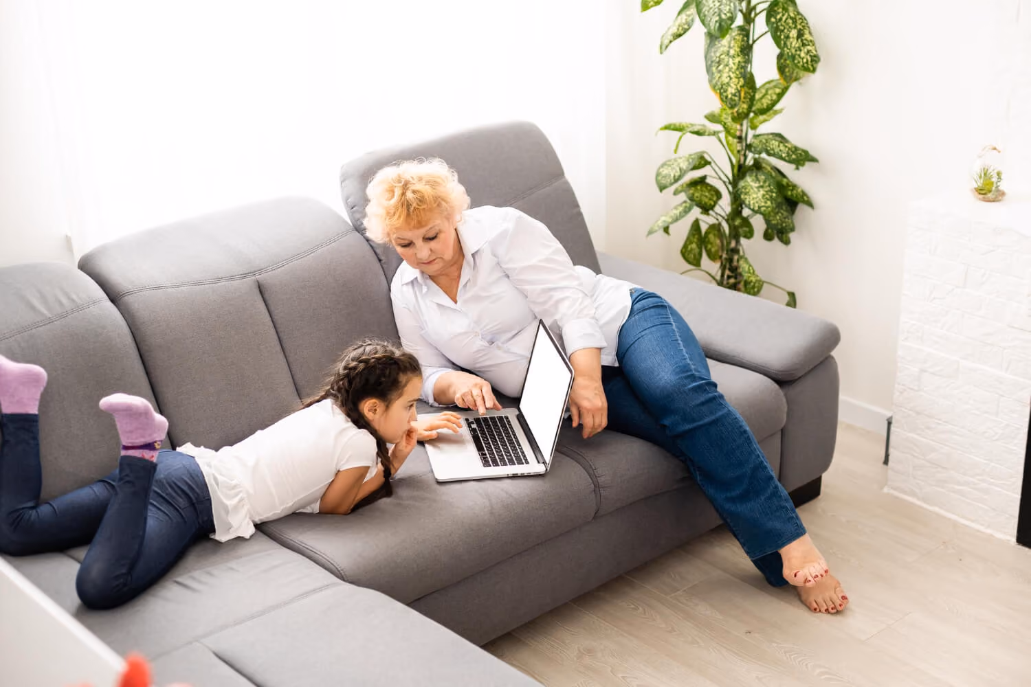 An elderly woman and a young girl lying on a gray sofa looking at a laptop together in a bright room with a green plant in the background.