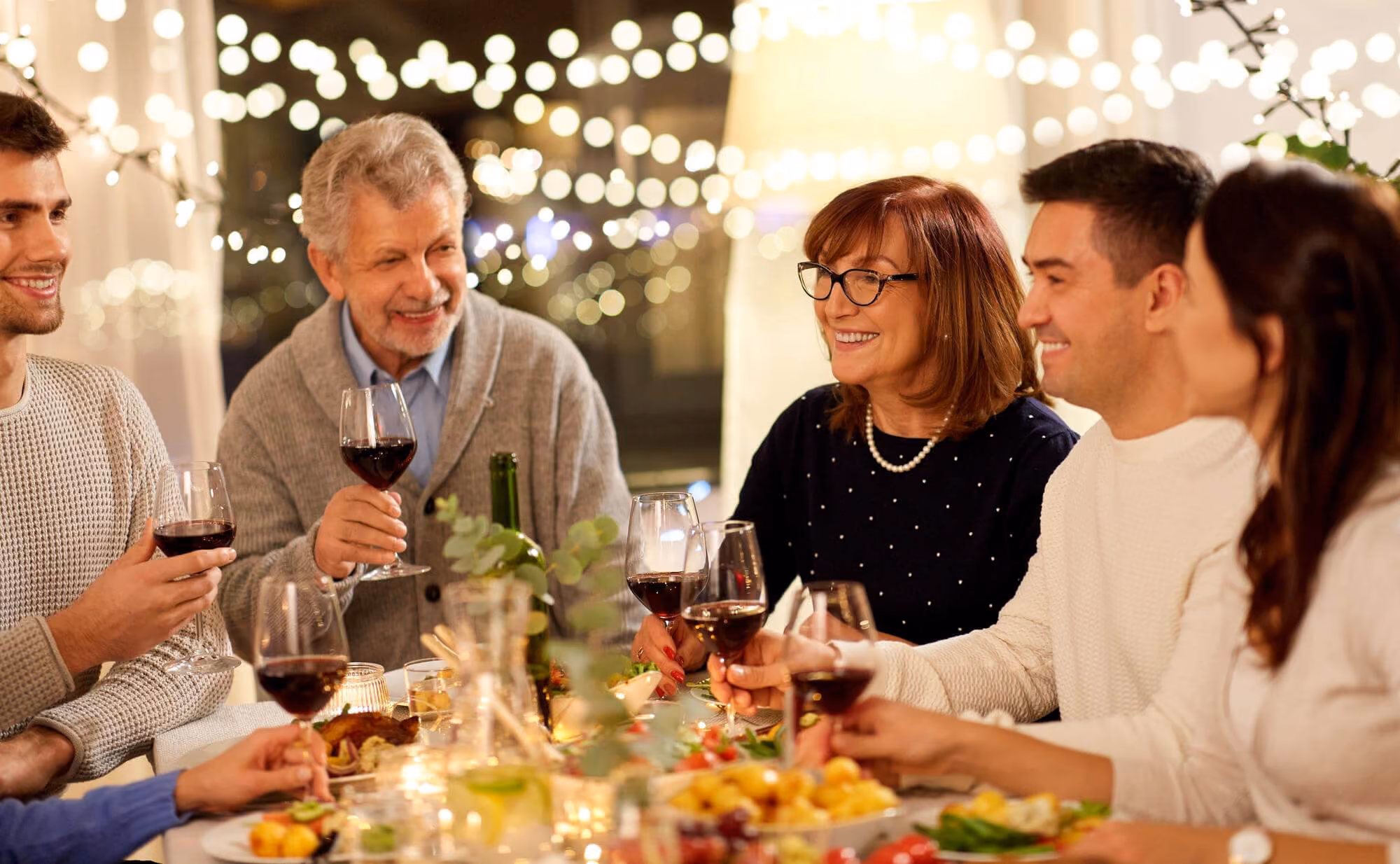 Five people sitting around a dinner table enjoying wine and food together, smiling and engaged in conversation under warm string lights.