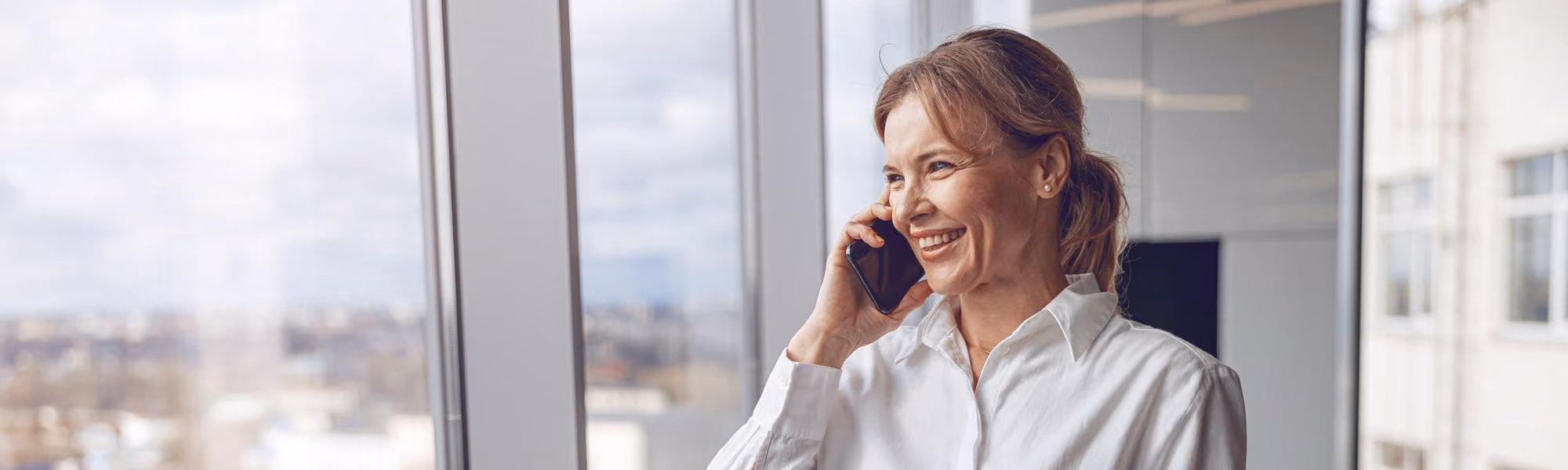 Smiling woman in a white shirt talking on a smartphone near a window with a city view in the background.