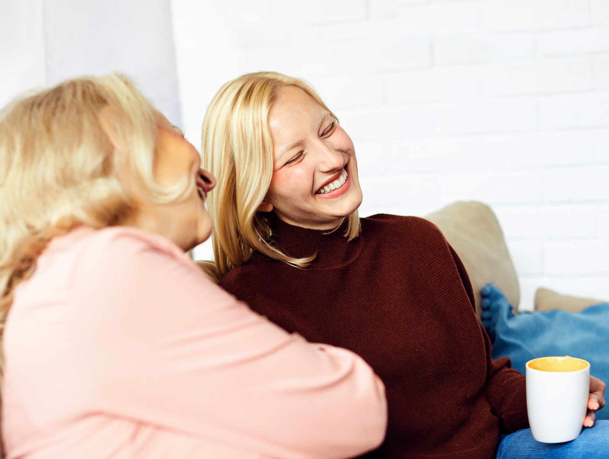 Two women sitting on a couch, smiling and engaged in conversation; one woman holds a white mug with a yellow interior.