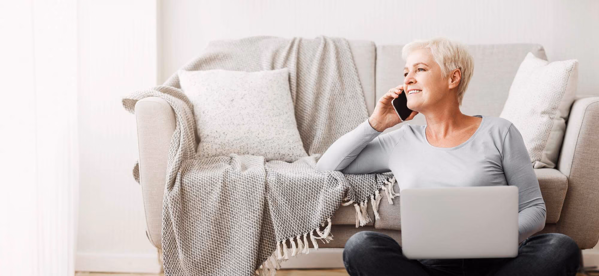 Smiling woman with short grey hair sitting on the floor in front of a beige sofa, holding a laptop on her lap and talking on a smartphone.