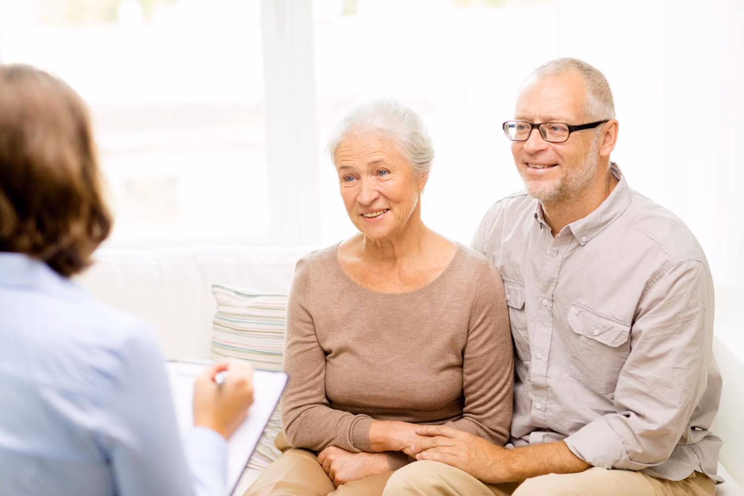 Smiling elderly woman and man sitting together on a couch, talking to a person holding a clipboard, suggesting a consultation or counseling session.