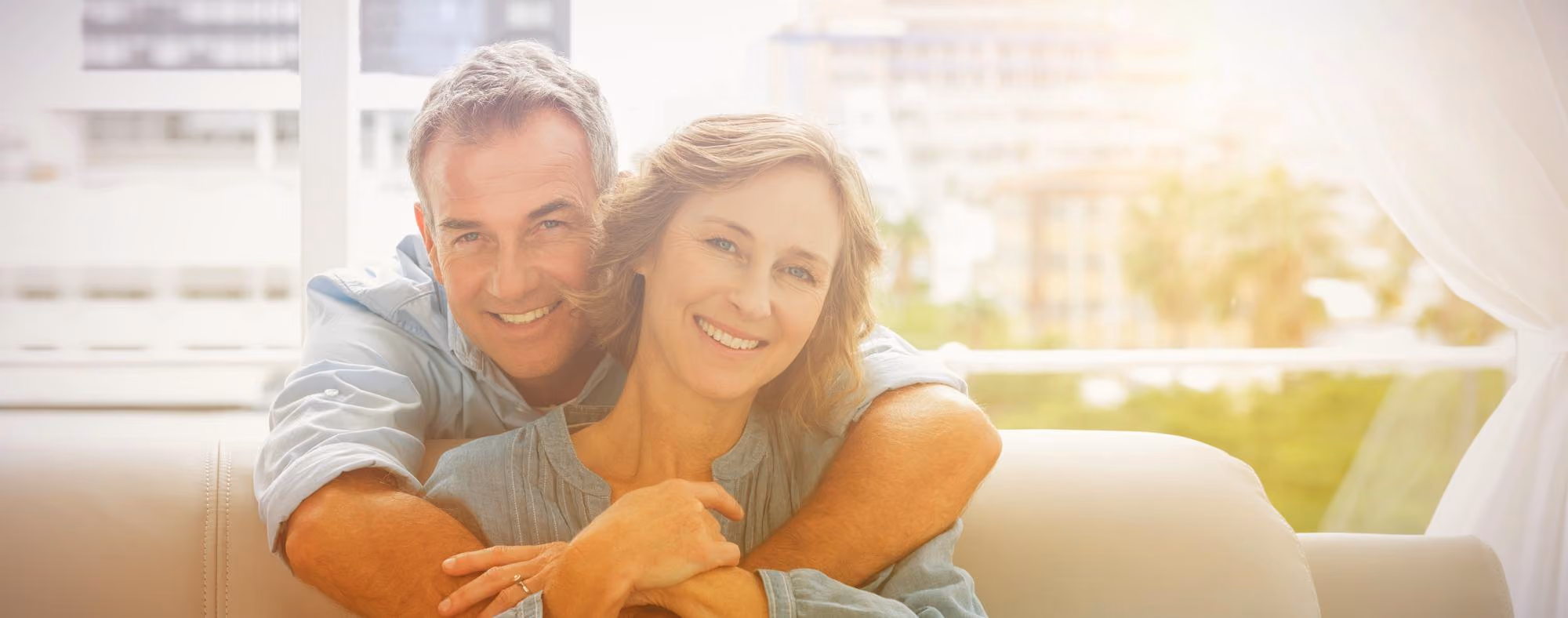 Smiling middle-aged man hugging a smiling middle-aged woman from behind on a sofa near a window with a bright, softly lit background.
