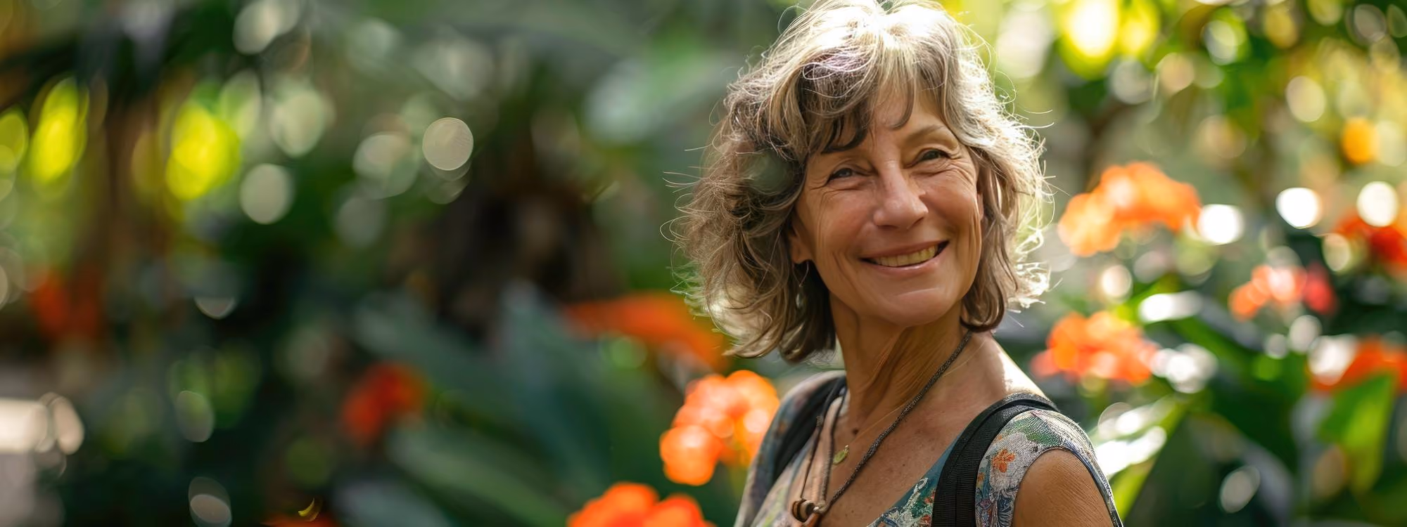 Smiling older woman with short wavy hair standing outdoors in front of blurred green foliage and orange flowers, wearing a patterned sleeveless top and necklace.