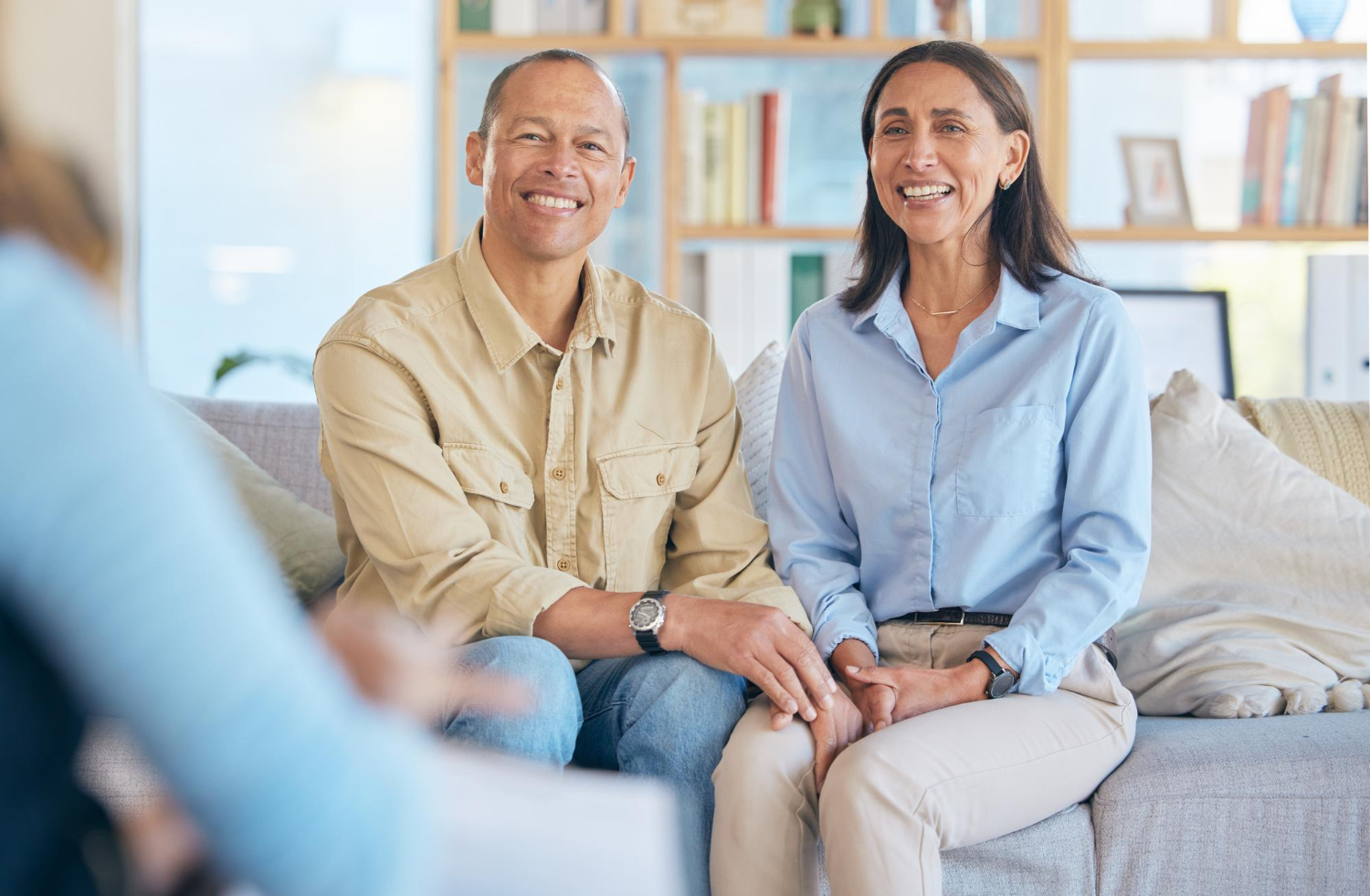 Smiling middle-aged couple sitting closely on a sofa, holding hands, with a blurred person in the foreground and a bookshelf in the background.