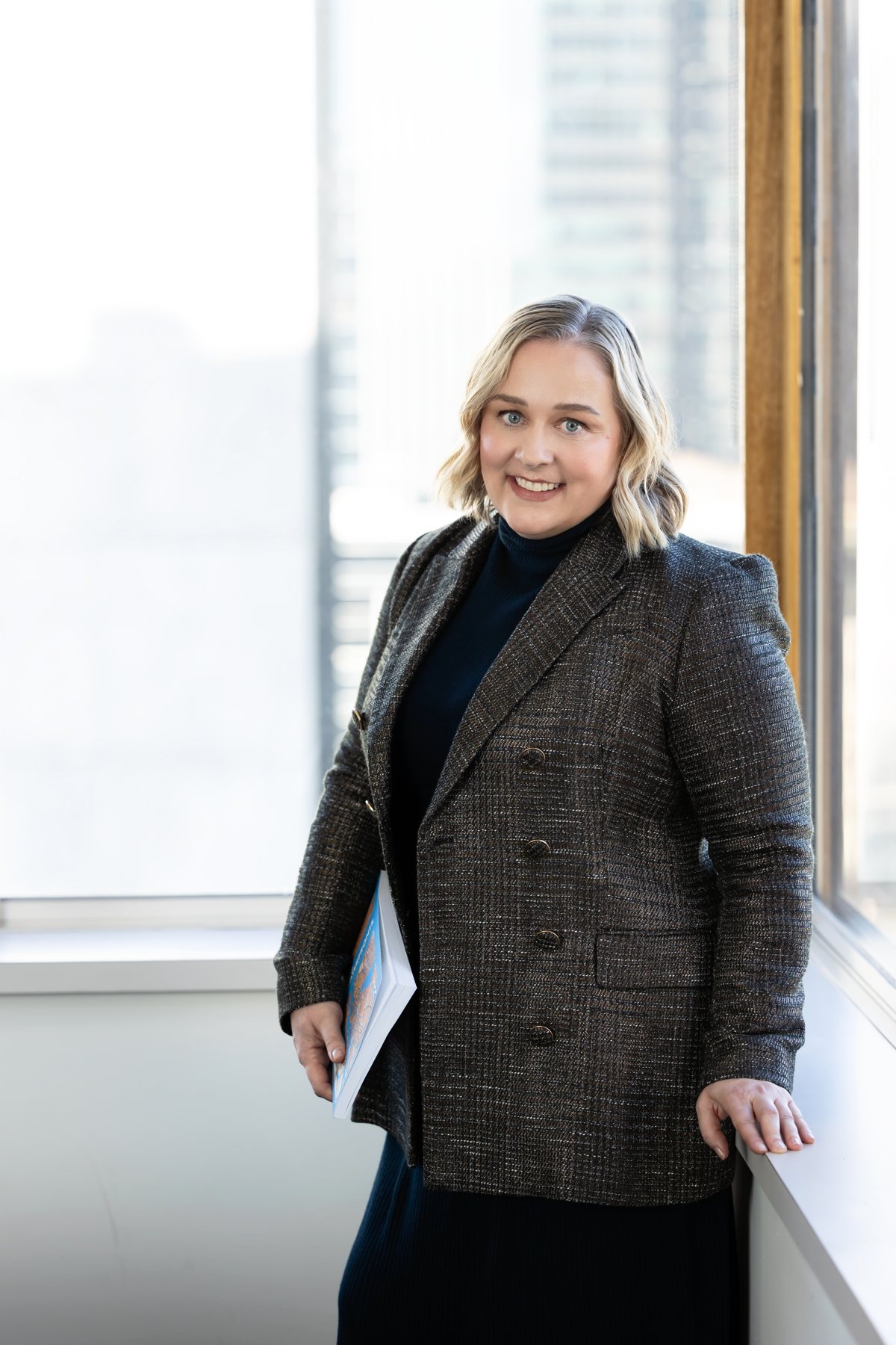 Catherine Hills smiling, standing by an office window in a tweed blazer, holding a book