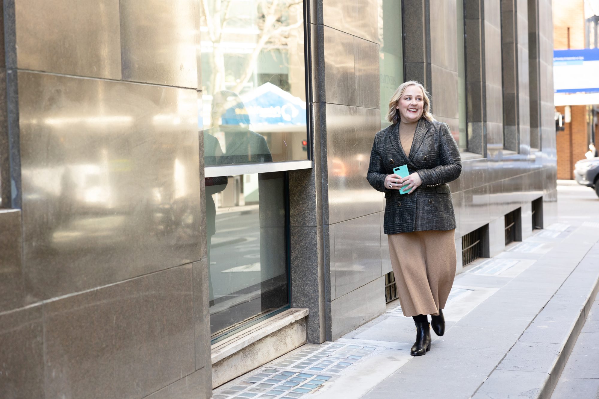 Catherine Hills smiling as she walks along a city street, holding her phone