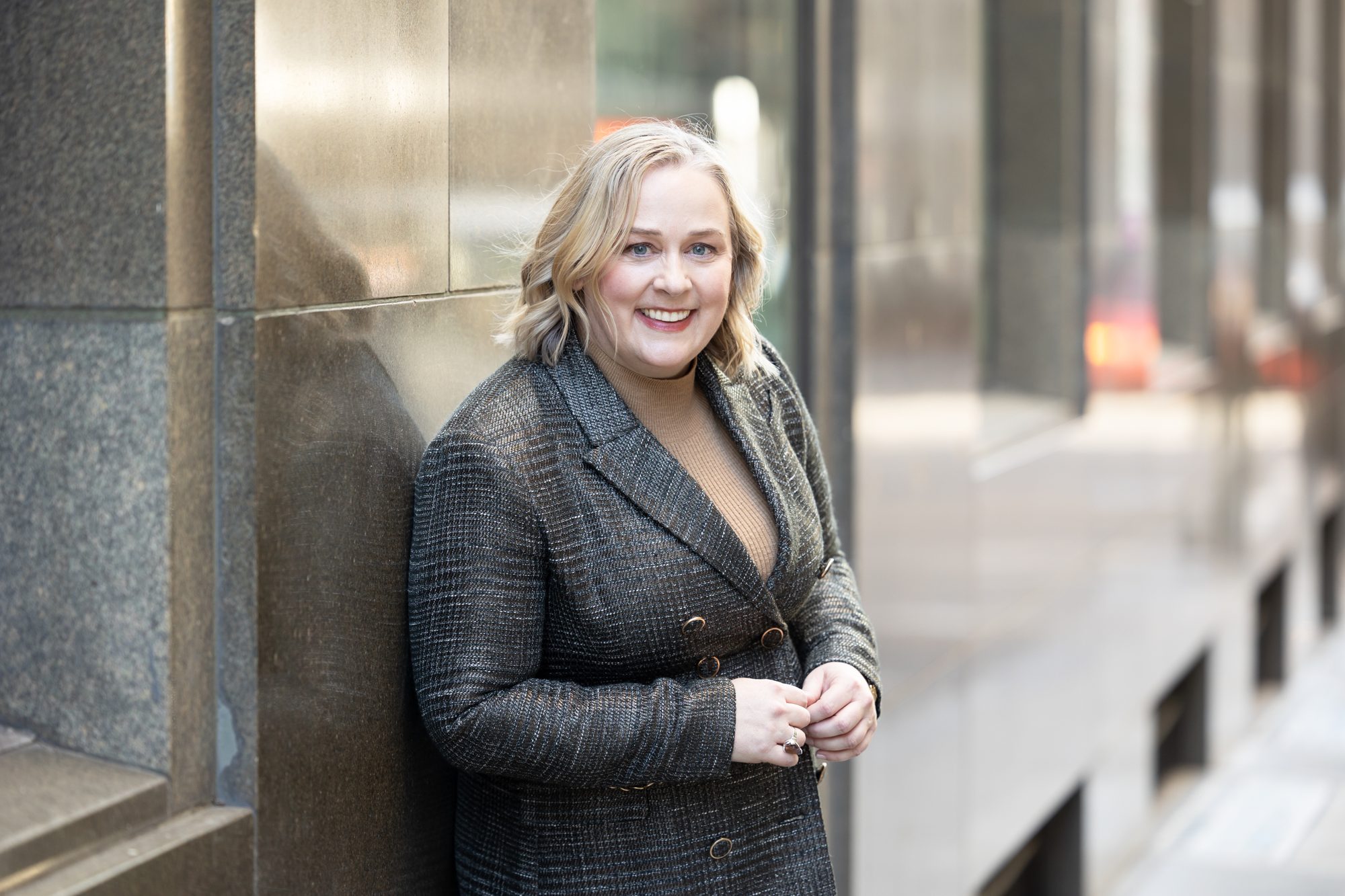 Catherine Hills smiling at the camera, leaning against a stone wall on a city street