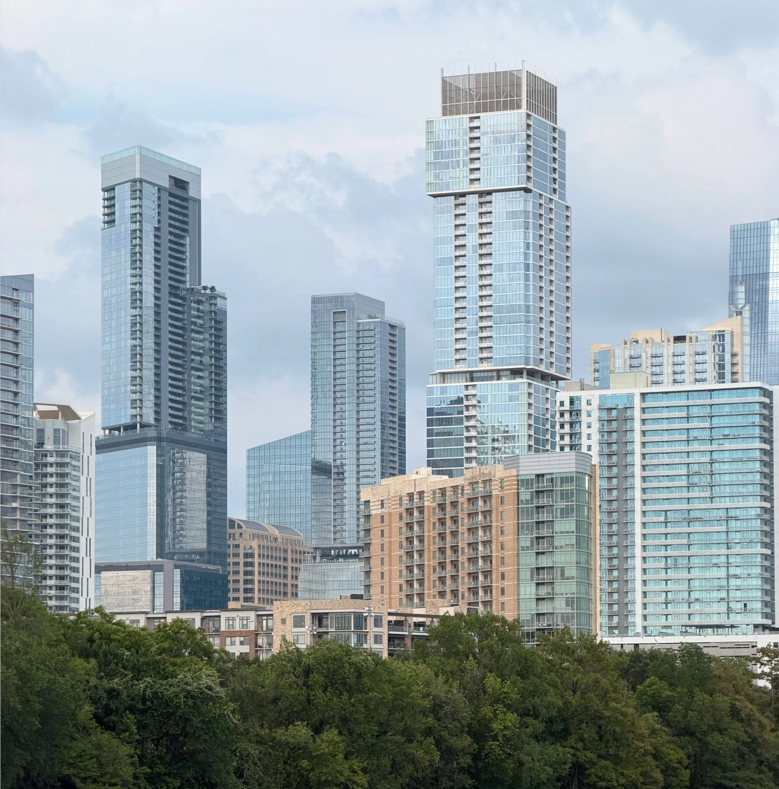City skyline with modern high-rise buildings behind dense green trees under a cloudy sky.