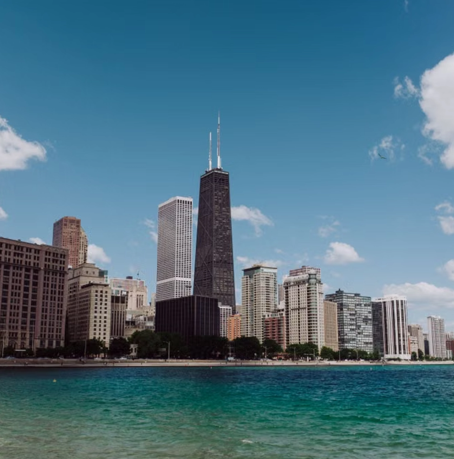 City skyline featuring tall buildings along a waterfront under a blue sky with scattered clouds.