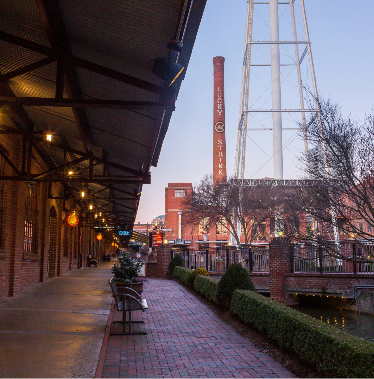 Brick walkway lined with benches and plants beside a building with illuminated hanging lights, leading toward a tall brick chimney with 'LUCKY STRIKE' written vertically, near a metal water tower and bare trees at dusk.