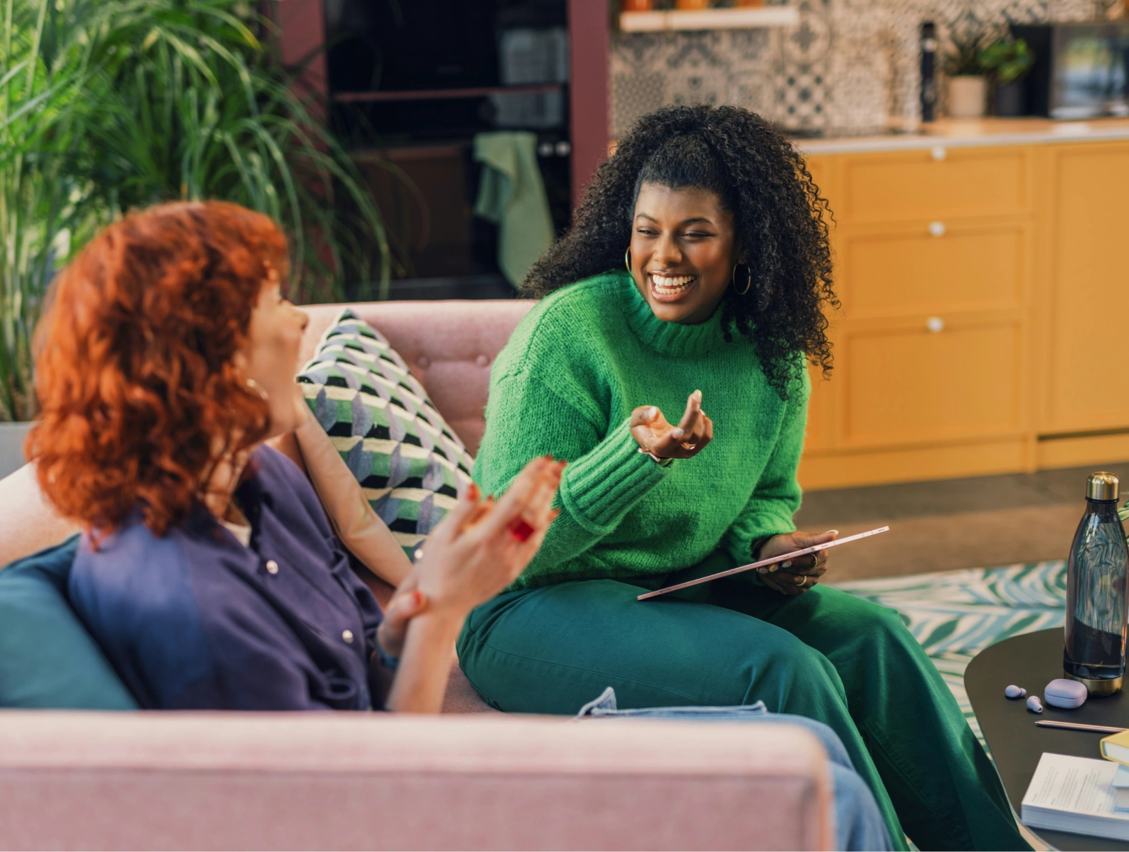 Two women sitting on a pink couch in a cozy room with plants and a kitchen in the background, smiling and conversing, one holding a tablet.