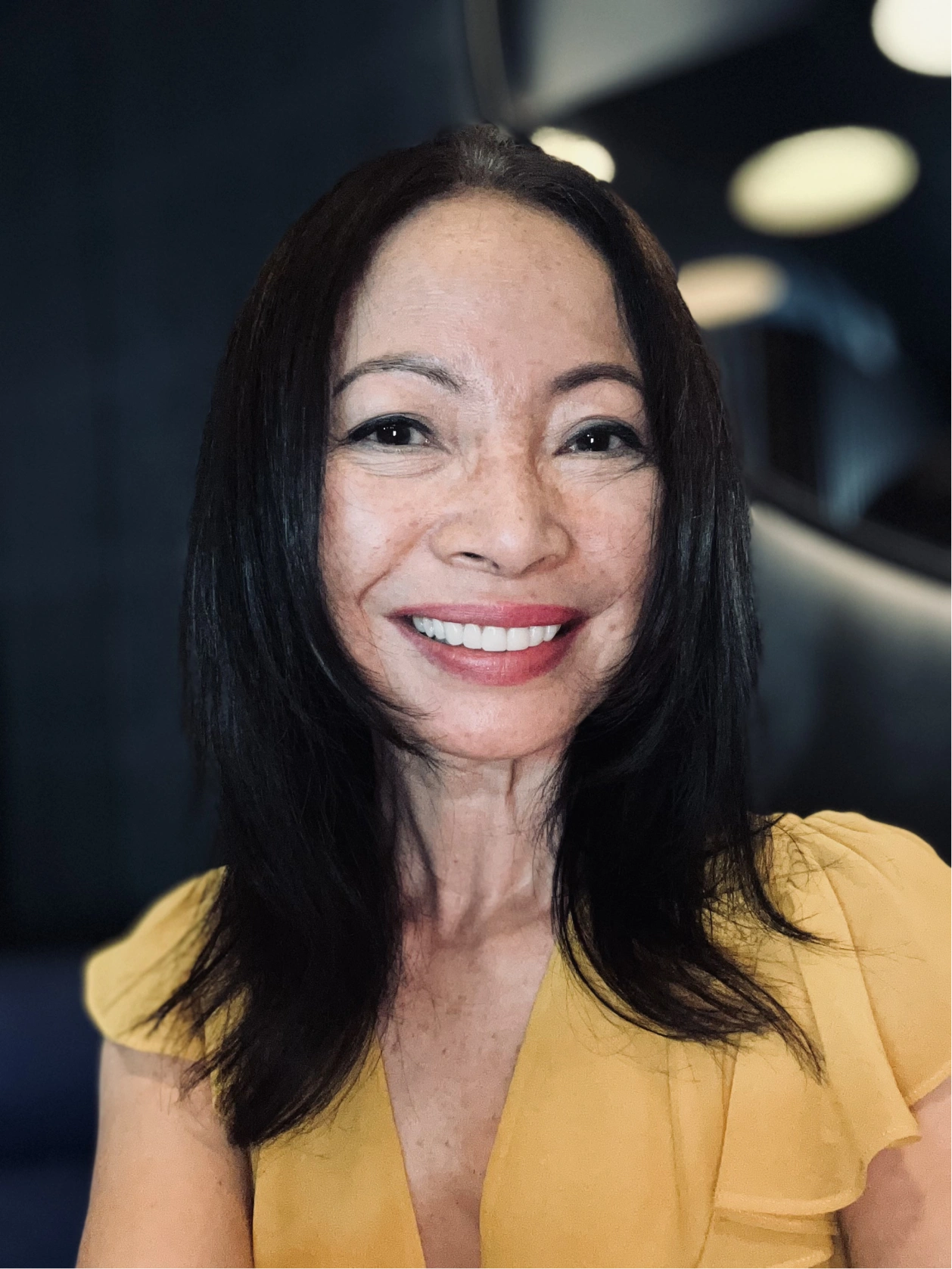 Smiling woman with long dark hair wearing a yellow top against a blurred dark background with round light spots.