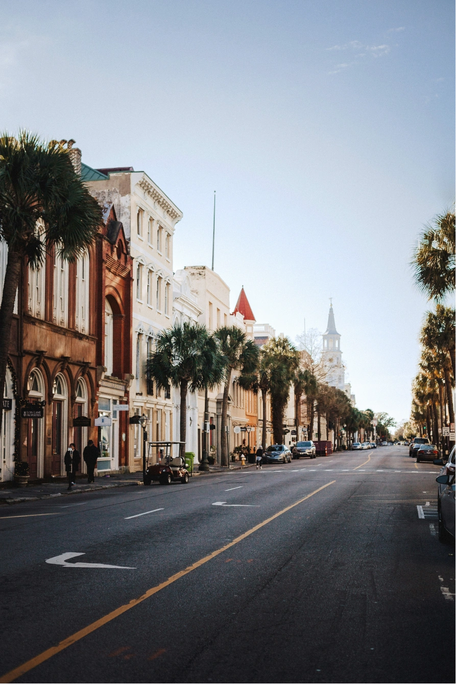 Urban street lined with historic buildings and palm trees under a clear blue sky with sparse clouds, few pedestrians on sidewalk, and cars parked along both sides of the street.
