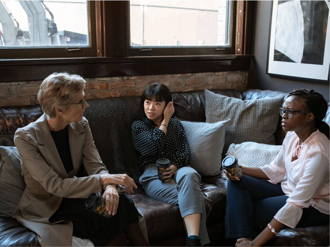 Three women sitting on a dark leather couch in a cozy room with large windows, having a conversation and holding coffee cups, surrounded by cushions.