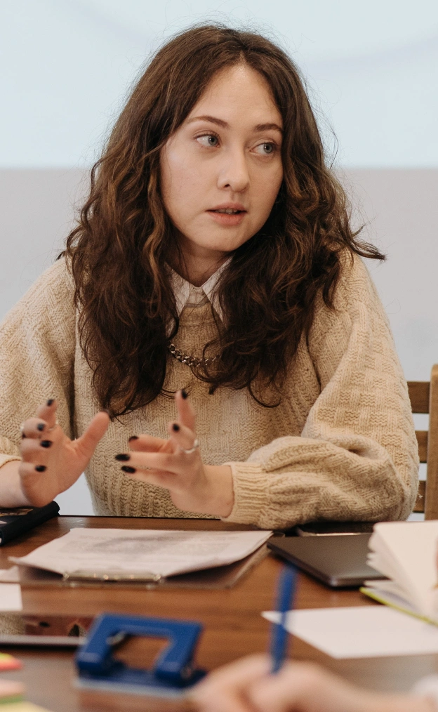 Young woman with curly brown hair wearing a beige cable-knit sweater and a gold chain necklace, sitting at a table with papers, speaking and gesturing with her hands.