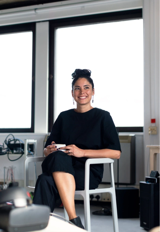 Smiling woman with dark hair in a bun, wearing a black outfit and sitting cross-legged on a gray chair in a modern office with large windows, holding a cup.