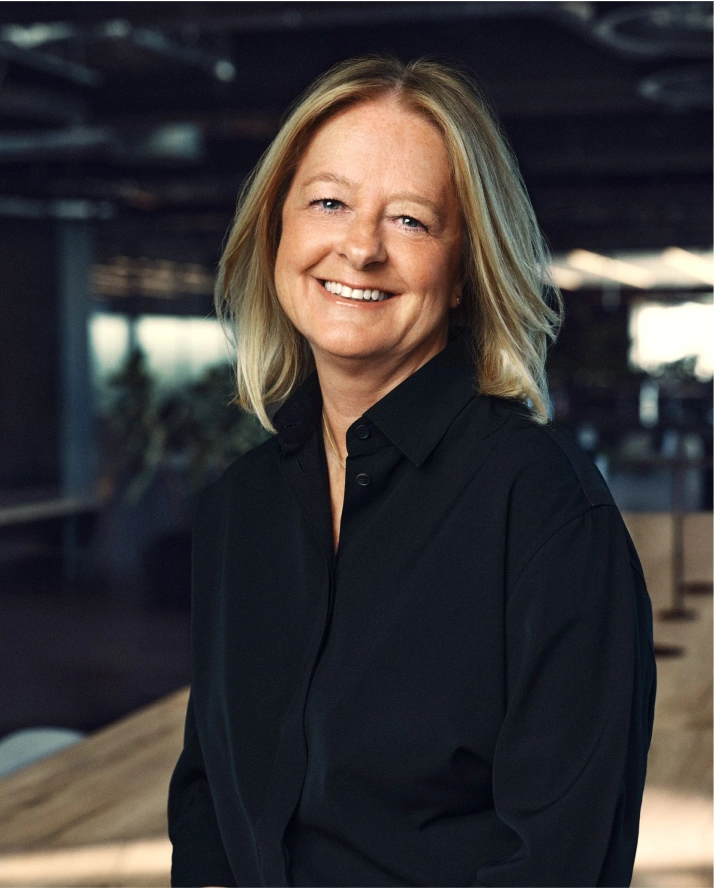 Smiling middle-aged woman with shoulder-length blonde hair wearing a black button-up shirt in a softly lit indoor space with blurred background.