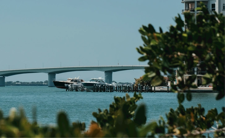 View of a calm blue waterway with boats docked at a pier, a bridge spanning across the water in the background, and green leafy plants framing the foreground.