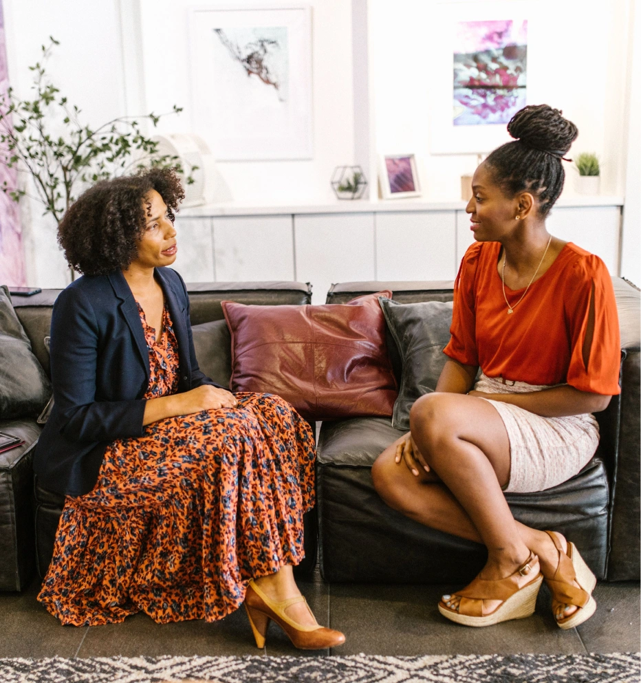 Two women sit on black leather sofas facing each other in a bright room, engaged in conversation, with decorative pillows and framed artwork behind them.