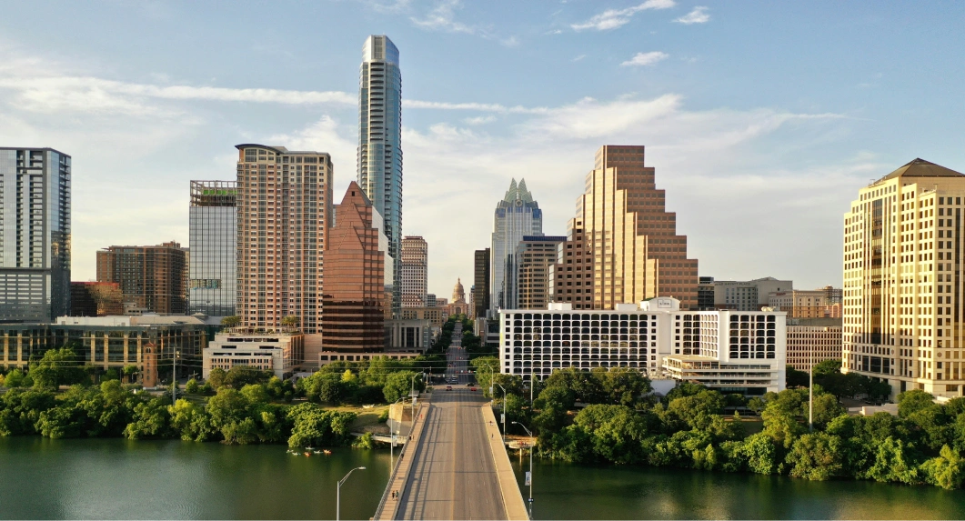City skyline with a mix of modern tall buildings and a stepped pyramid-shaped skyscraper by a river, connected by a bridge lined with trees on both sides under a partly cloudy sky.