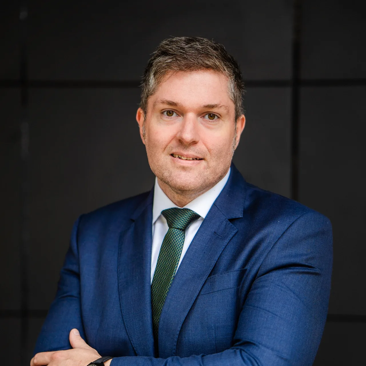 James Walker in a navy suit and green tie, arms folded, against a dark panelled wall