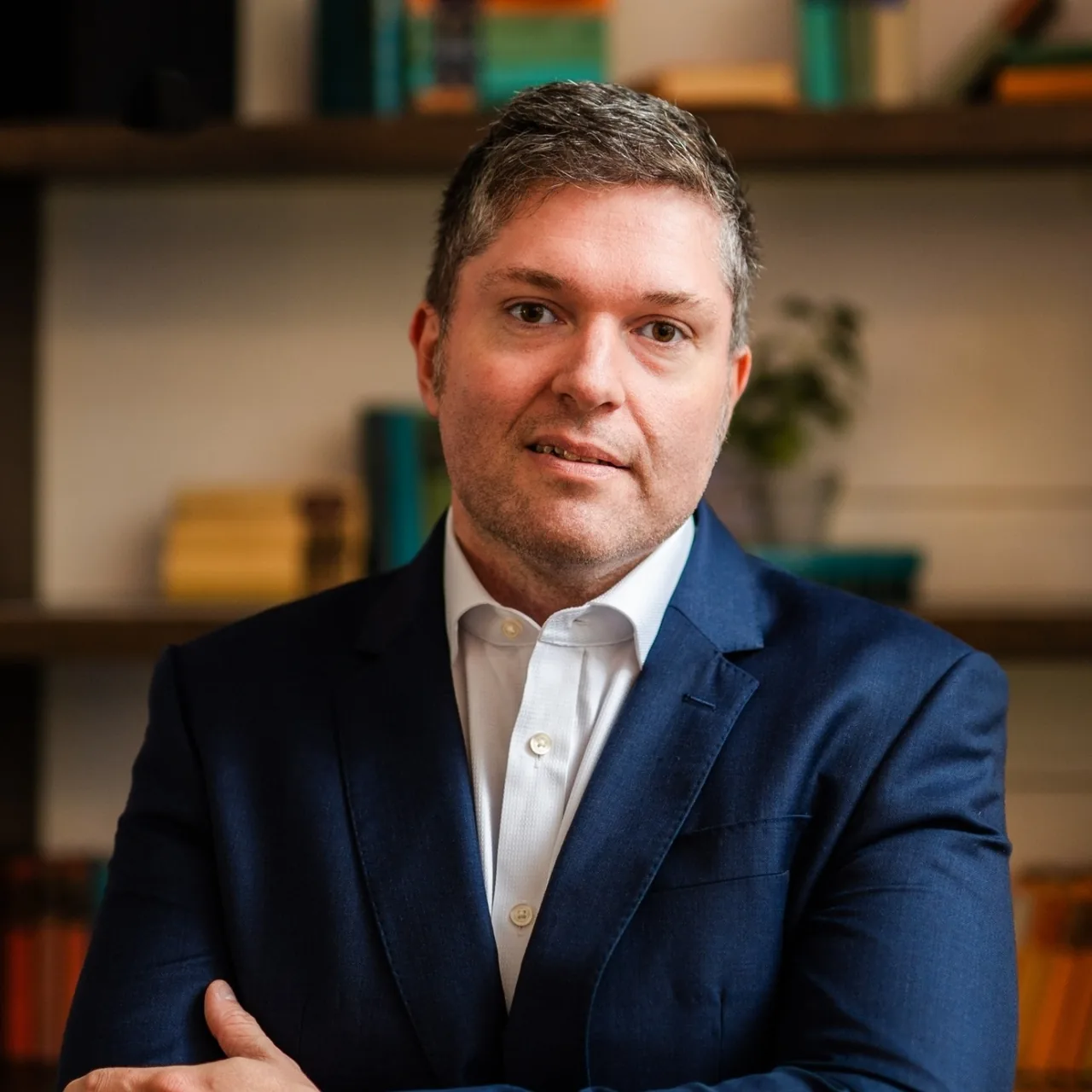 James Walker in a navy jacket and open-necked white shirt, in front of bookshelves