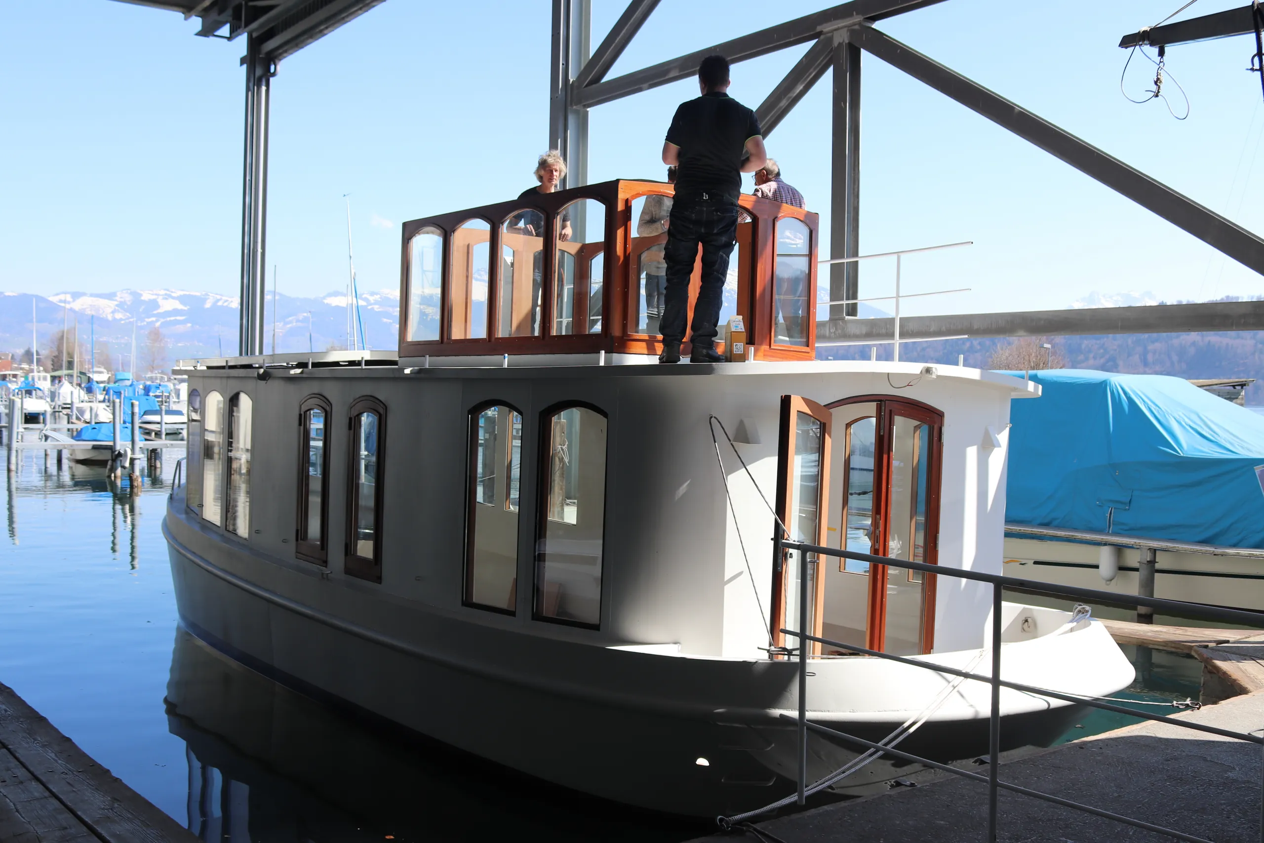 White boat docked at a marina with wooden framed windows and an upper deck cabin with people standing inside and on the deck, with a backdrop of snow-capped mountains and other boats.