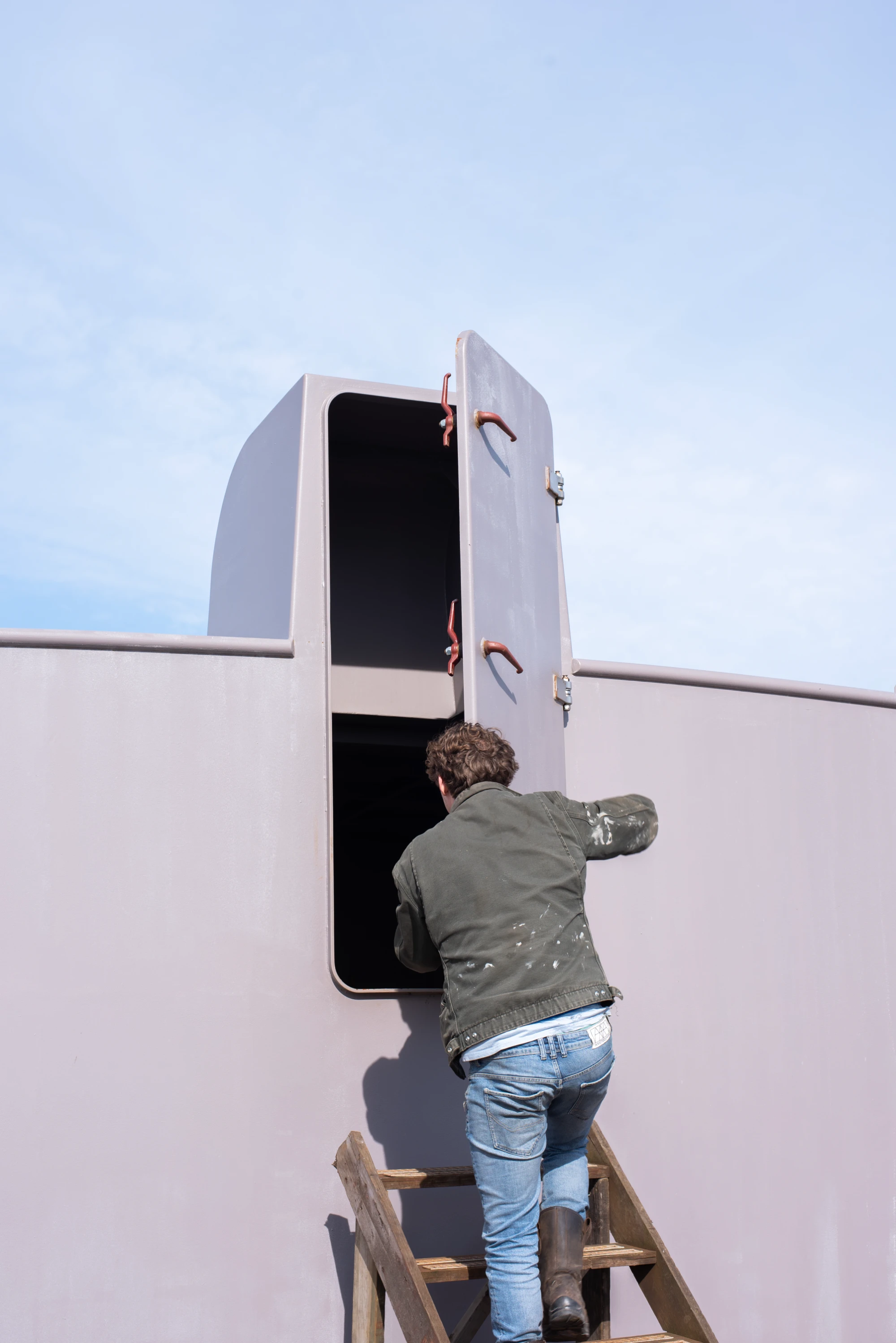 Person wearing a dark jacket, light blue jeans, and boots climbing wooden stairs and entering an open hatch door on a large gray structure against a blue sky.