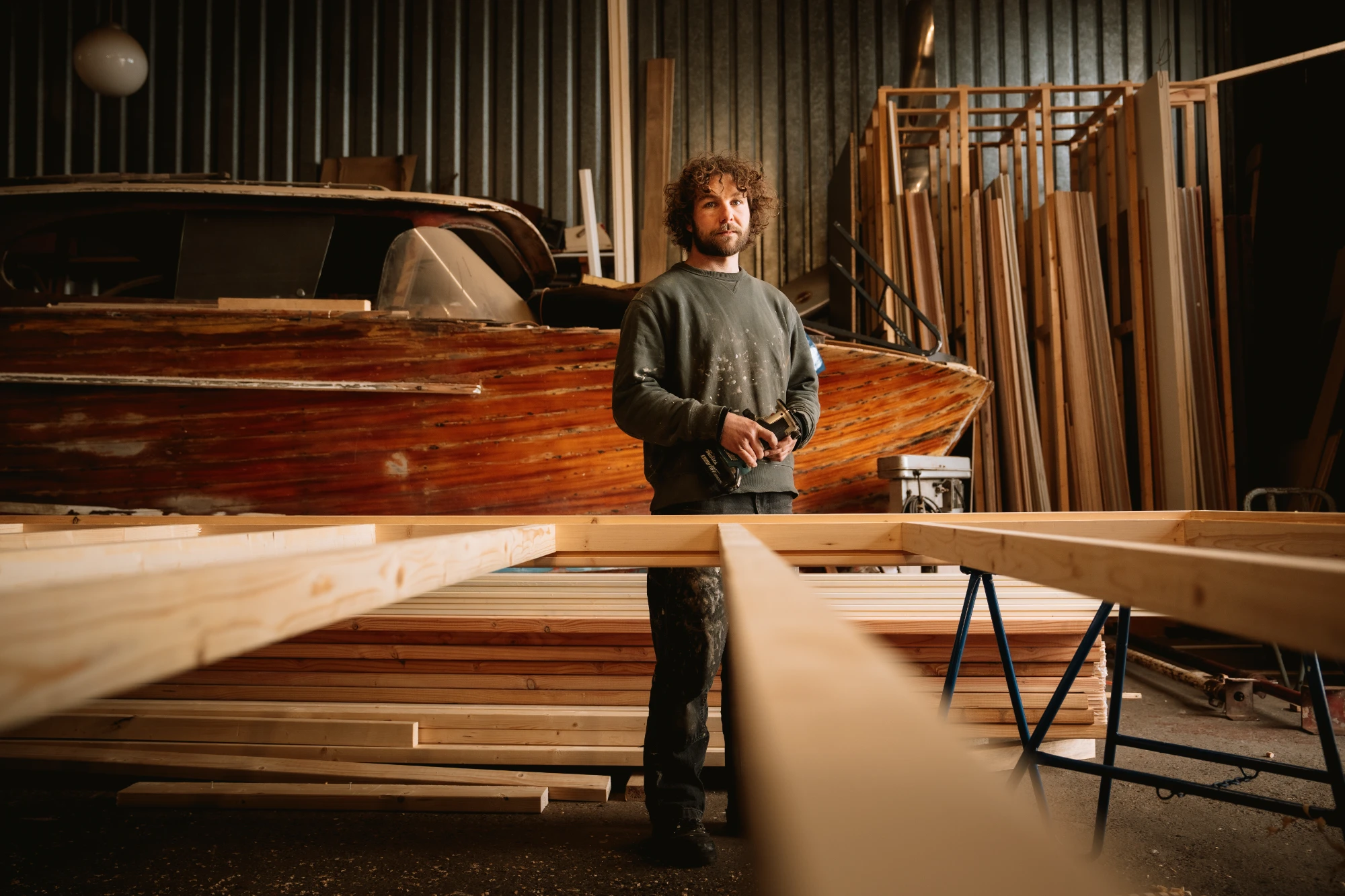 A man with curly hair and a beard stands in a workshop holding a power tool, with wooden planks and a large wooden boat in the background.
