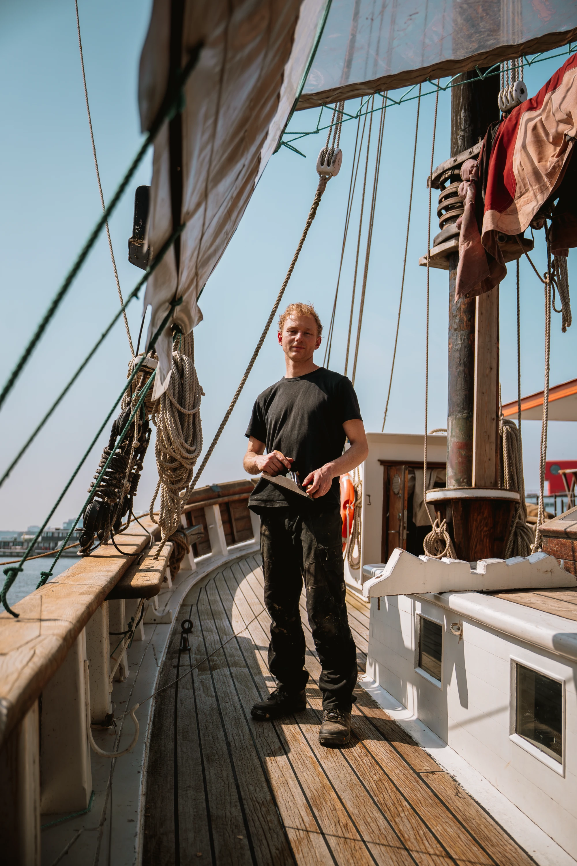 Man with blonde hair wearing a black t-shirt and pants standing on the wooden deck of a sailboat, holding a tool, surrounded by ropes and sails under a clear sky.