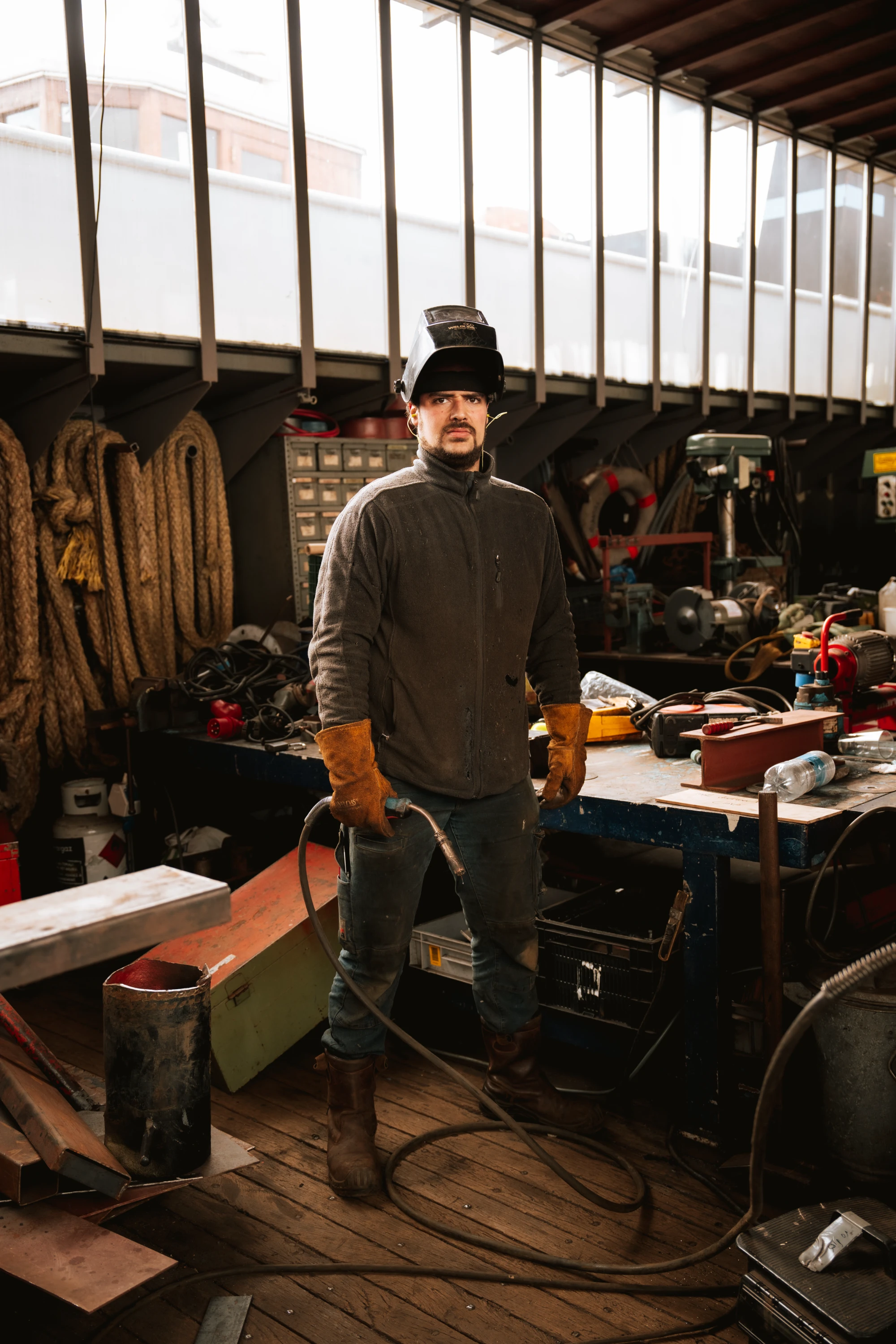 Man in a workshop wearing a welding helmet lifted up, brown gloves, and work boots, holding a welding torch connected by a hose, standing on wooden floor near a cluttered workbench with tools and equipment.