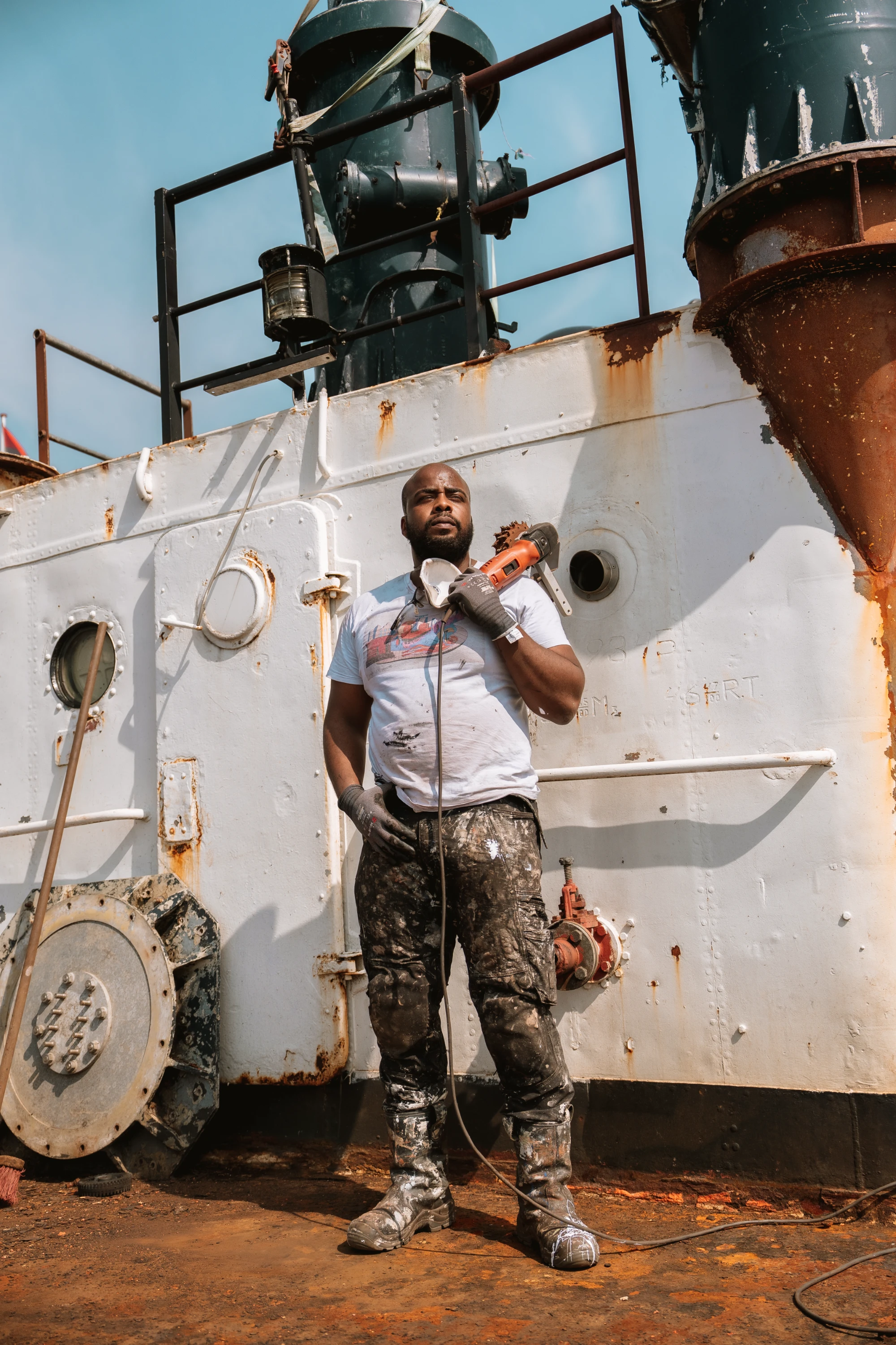 Man in work clothes standing on a rusty metal surface in front of a worn white industrial structure, holding an orange power tool over his shoulder and wearing gloves and boots covered in paint and dirt.