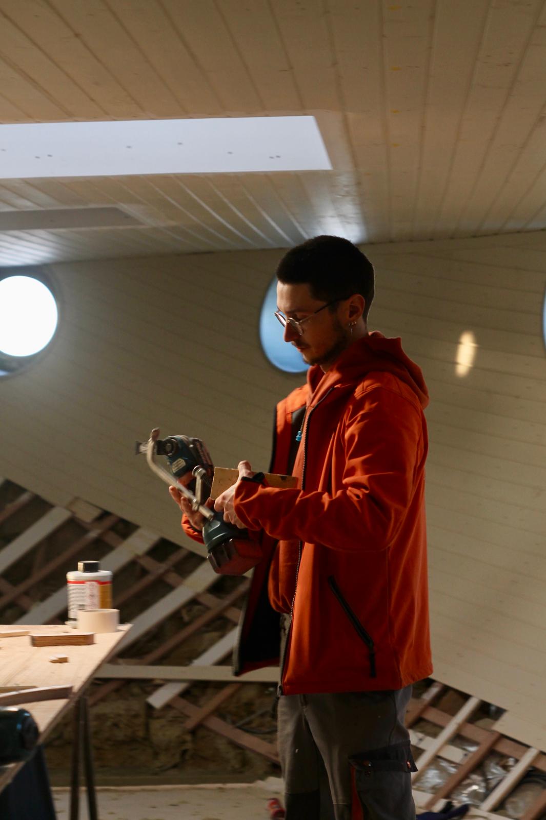 Man wearing an orange jacket holding a power drill while working indoors near a wooden workbench with tools and materials.