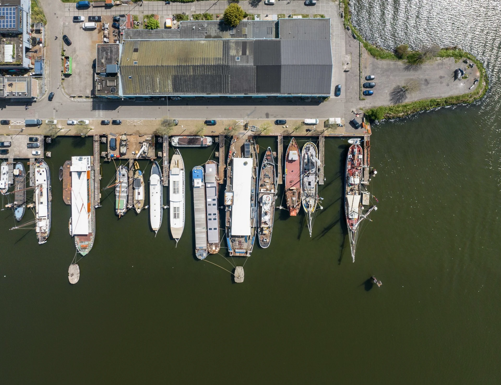 Aerial view of a dock with multiple boats and ships moored side by side alongside a building with a segmented roof, surrounded by water and parked cars.