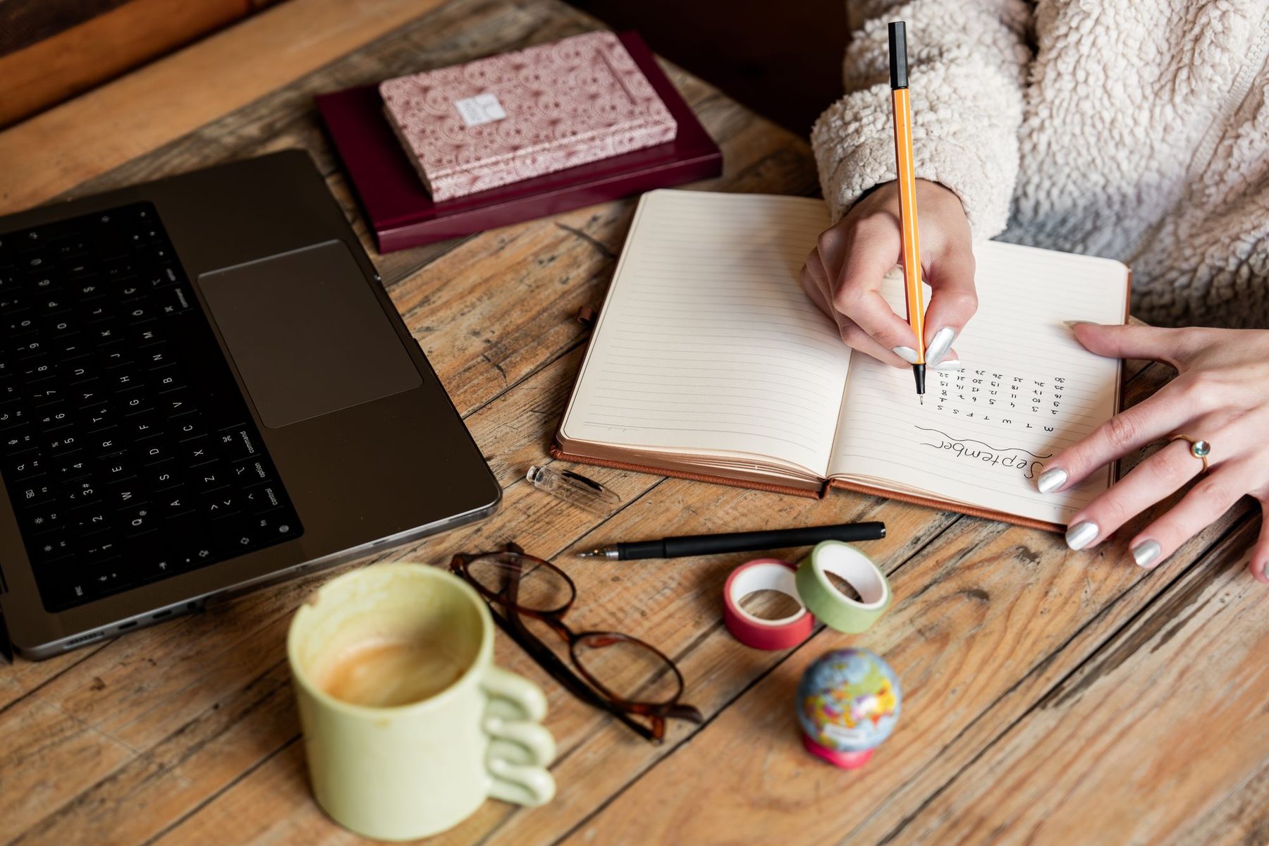Hands writing title ideas in a notebook beside a laptop and coffee mug