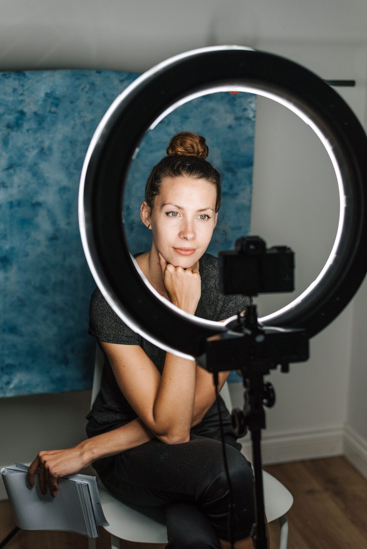 Presenter seated inside a ring light facing a camera on a stand in front of a blue backdrop