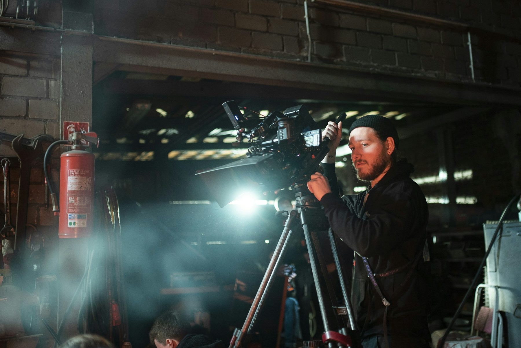 Camera operator filming on a tripod in a hazy warehouse set with strong backlight