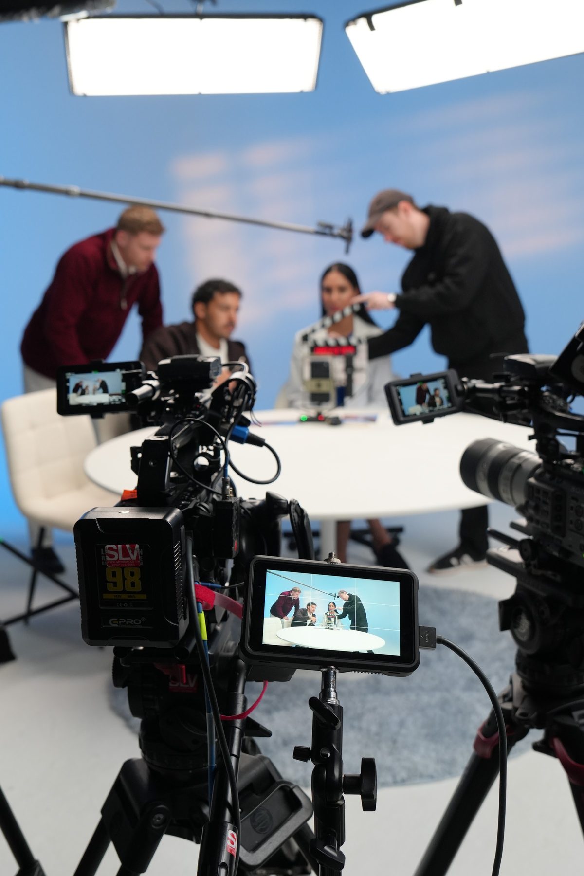 Crew preparing presenters at a round table on a bright studio set with cameras in the foreground